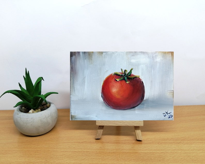 May include: A small painting of a red tomato on a white background. The painting is on a small easel.