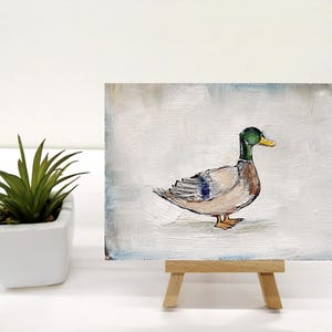 May include: A small painting of a duck on a white background. The duck is brown with a green head and a yellow beak. The painting is on a small wooden easel. The artist's signature is in the bottom right corner of the painting. Mallard duck painting original miniature art Vintage duck oil painting farmhouse kitchen wall art country decor duck hunting gift 4x6