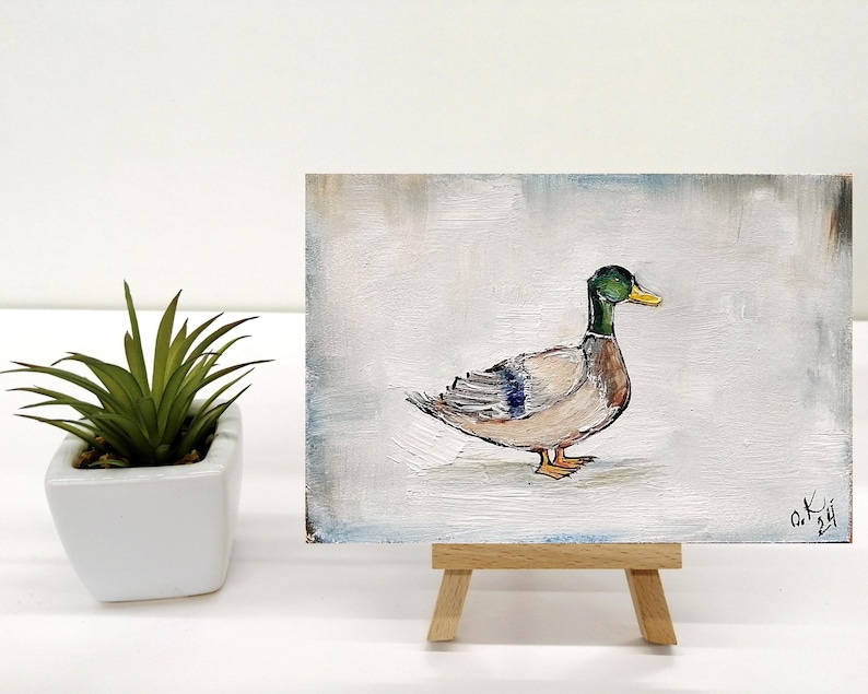 May include: A small painting of a duck on a white background. The duck is brown with a green head and a yellow beak. The painting is on a small wooden easel. The artist's signature is in the bottom right corner of the painting. Mallard duck painting original miniature art Vintage duck oil painting farmhouse kitchen wall art country decor duck hunting gift 4x6