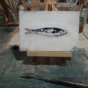 May include: An original oil painting of a sardine on a small white canvas. The fish is depicted in shades of silver, black, and white, with a textured white background. The artwork is displayed on a miniature wooden easel.