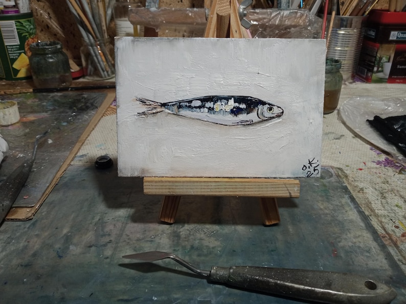 May include: An original oil painting of a sardine on a small white canvas. The fish is depicted in shades of silver, black, and white, with a textured white background. The artwork is displayed on a miniature wooden easel.