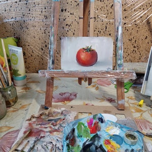 May include: A small painting of a red tomato on a white background, displayed on a wooden easel. The painting is surrounded by art supplies, including paintbrushes, a palette, and a jar of paint.