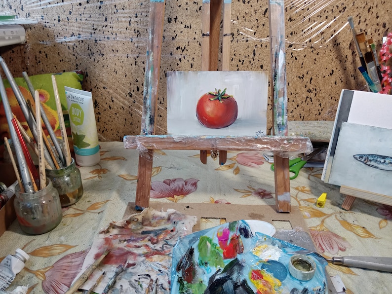 May include: A small painting of a red tomato on a white background, displayed on a wooden easel. The painting is surrounded by art supplies, including paintbrushes, a palette, and a jar of paint.