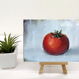May include: A small painting of a red tomato on a white background. The tomato is painted in a realistic style with a green stem and leaves. The painting is on a small easel.