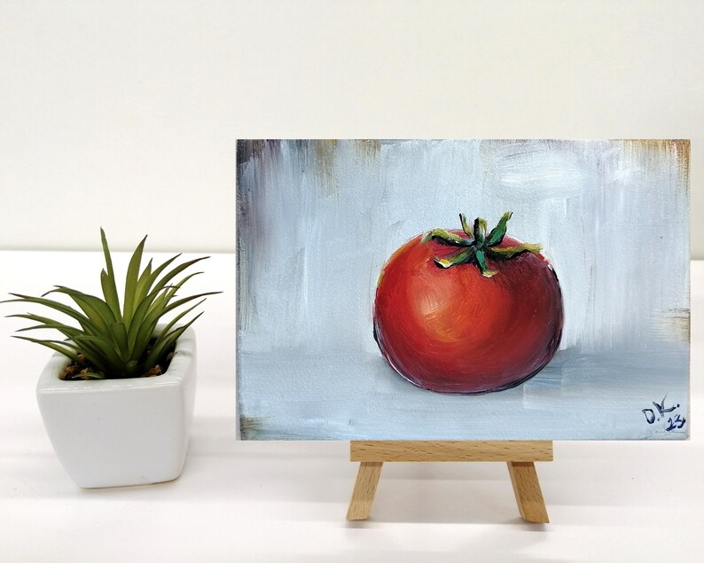 May include: A small painting of a red tomato on a white background. The tomato is painted in a realistic style with a green stem and leaves. The painting is on a small easel.