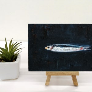 May include: A small painting of a silver fish on a black background. The fish has a red stripe on its side. The painting is on a small easel. The artist's signature and the year 2023 are in the bottom right corner of the painting. Sardine art original painting Sardines still life oil painting miniature Anchovy fish painting French country kitchen art 4x6