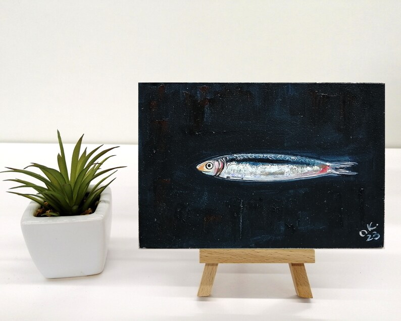 May include: A small painting of a silver fish on a black background. The fish has a red stripe on its side. The painting is on a small easel. The artist's signature and the year 2023 are in the bottom right corner of the painting. Sardine art original painting Sardines still life oil painting miniature Anchovy fish painting French country kitchen art 4x6