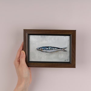 May include: A framed painting of a single, silver-colored fish with blue and black markings on a white background. The frame is dark brown wood.