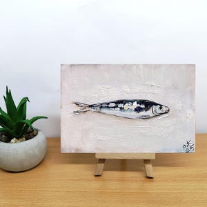May include: Oil painting of a sardine on a small easel. The fish is painted in shades of black, white, and silver against a textured white background. A small potted plant sits to the left of the painting.
