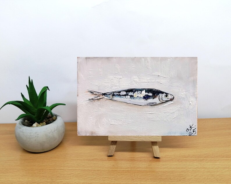 May include: Oil painting of a sardine on a small easel. The fish is painted in shades of black, white, and silver against a textured white background. A small potted plant sits to the left of the painting.