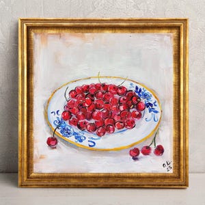 May include: An oil painting in a gold frame depicting a white plate with blue floral patterns filled with bright red cherries. Several loose cherries are scattered near the plate on a light, textured background. The artist's signature O.K. 23 is visible.