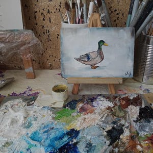 May include: A small painting of a duck on a wooden easel. The duck is painted in shades of brown, gray, and green. The background of the painting is a light blue color. The painting is on a table with a paint palette and a small container of paint. Mallard duck painting original miniature art Vintage duck oil painting farmhouse kitchen wall art country decor duck hunting gift 4x6