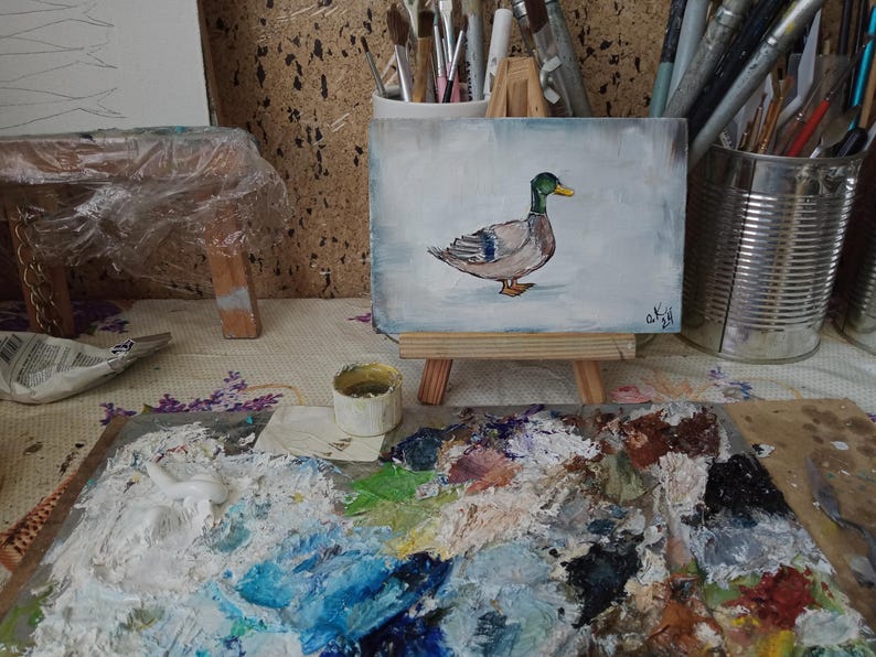 May include: A small painting of a duck on a wooden easel. The duck is painted in shades of brown, gray, and green. The background of the painting is a light blue color. The painting is on a table with a paint palette and a small container of paint. Mallard duck painting original miniature art Vintage duck oil painting farmhouse kitchen wall art country decor duck hunting gift 4x6