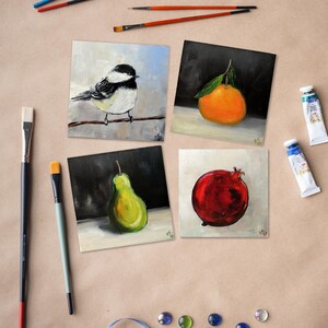 May include: Four square paintings of fruit and a bird, with paint tubes and brushes. The paintings depict a bird on a branch, an orange with a leaf, a green pear, and a red pomegranate. The art supplies are scattered around the paintings.