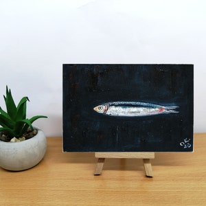 May include: A small painting of a silver fish on a black background. The fish is facing to the right and has a red tail fin. The painting is signed 'OK 23' in the bottom right corner. Sardine art original painting Sardines still life oil painting miniature Anchovy fish painting French country kitchen art 4x6