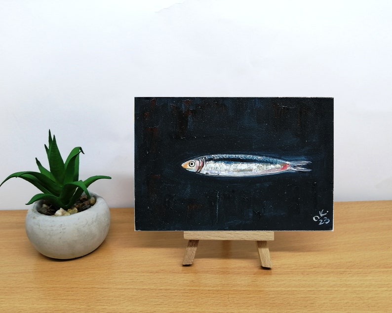 May include: A small painting of a silver fish on a black background. The fish is facing to the right and has a red tail fin. The painting is signed 'OK 23' in the bottom right corner. Sardine art original painting Sardines still life oil painting miniature Anchovy fish painting French country kitchen art 4x6
