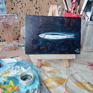 May include: A small painting of a silver fish on a black background. The painting is on a wooden easel and has the signature 'OK. 23' in the bottom right corner. Sardine art original painting Sardines still life oil painting miniature Anchovy fish painting French country kitchen art 4x6