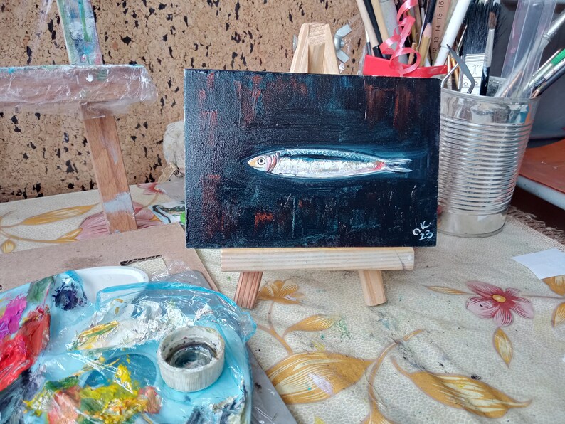 May include: A small painting of a silver fish on a black background. The painting is on a wooden easel and has the signature 'OK. 23' in the bottom right corner. Sardine art original painting Sardines still life oil painting miniature Anchovy fish painting French country kitchen art 4x6