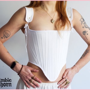 May include: A white cotton corset with a pointed bottom and adjustable straps. The corset has a ribbed texture and is worn by a person with tattoos on their arms.