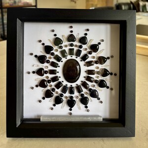 May include: A square black frame containing a white background with a circular mandala arrangement of dark brown and black gemstones. The design features heart-shaped stones, faceted rectangular pieces, and small clear crystal points.