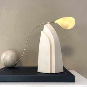 May include: A white ceramic sculpture with a curved design, a gold metal stem with a leaf-shaped top, and a round gray stone on a black book.