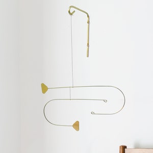 May include: A brass-coloured mobile with a minimalist design hangs against a white wall. The mobile features curved lines and geometric shapes, including two heart-shaped elements. The design is suspended by a thin wire, adding a touch of modern art to the room.