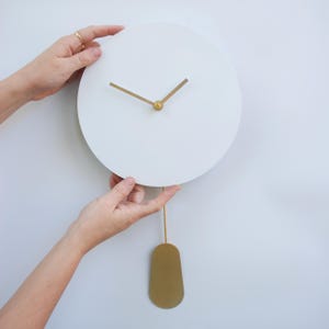 May include: A white, circular wall clock with gold hands and a gold pendulum. The clock is held by two hands against a white background. The clock's minimalist design is modern and elegant.