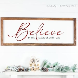 Puede incluir: Un letrero de madera con el texto "Believe in the magic of Christmas" en letras cursivas rojas. El letrero está colgado en una pared blanca con una repisa blanca debajo. La repisa está decorada con una pequeña figurita de casa blanca y una rama de vegetación escarchada.