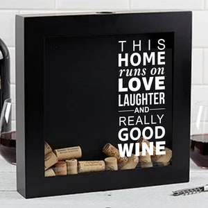 May include: Black wooden box with a glass front, displaying a collection of wine corks. The box has a white inscription that reads "This home runs on love, laughter, and really good wine."