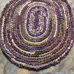 May include: Oval braided rug in shades of purple, brown, and cream. The rug is made of fabric strips sewn together in a spiral pattern. The rug's design features a mix of solid colors and plaid patterns.