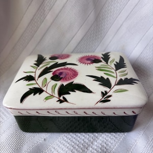 May include: A square ceramic trinket box with a white lid featuring a floral design with pink and green accents. The box has a dark green base and a decorative border. The lid is adorned with stylized thistles.