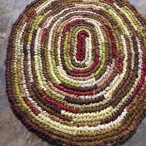 May include: A handmade, circular rag rug with a spiral design. The rug features a mix of brown, green, red, and cream-colored fabric strips. The center is an oval shape in a deep red color. The rug is approximately 36 inches in diameter.
