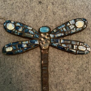 May include: A mosaic dragonfly ornament with blue and white wings. The wings are made of small, irregular tiles in shades of blue, with white accents. The body is a mix of silver and brown tones, and the head is a bright blue oval.