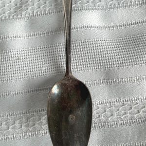 May include: A tarnished silver-toned metal spoon featuring a slender handle that widens toward the top. The bowl of the spoon is oval-shaped and shows visible signs of oxidation and wear, with a dark, mottled patina across its surface.