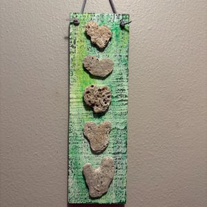 May include: A rectangular wooden wall hanging painted in shades of green with a distressed finish. Five heart-shaped stones are arranged vertically in a single column down the center of the board. A thin gray cord is attached for hanging purposes.