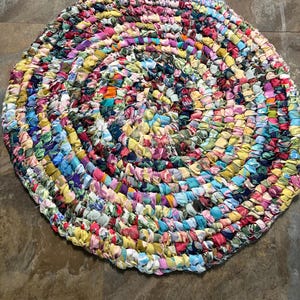 May include: A round, handmade rug with a spiral design. The rug is made of colorful fabric strips in various shades of pink, yellow, blue, and green. The rug's diameter is approximately 36 inches.
