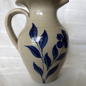 May include: A beige ceramic pitcher with a glossy finish, featuring a hand-painted dark blue floral design with leaves and a single flower. The pitcher has a curved handle on the left side and a wide, flared spout at the top for pouring liquids.