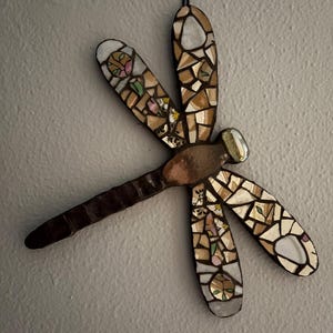 May include: A mosaic dragonfly wall hanging. The dragonfly features brown, tan, and white mosaic tiles on the wings, with a dark brown body. The wings have a mix of shapes and colors, with some sections featuring floral designs. A wire is attached for hanging.