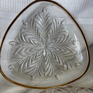 May include: A triangular clear glass dish featuring a pressed leaf or floral pattern radiating from the center. The edges of the dish are finished with a thin gold-colored metallic trim, creating a distinct border around the glass form.