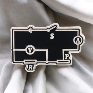 May include: A black and silver enamel pin featuring a circuit diagram design. The pin includes the letters "V", "A", "R", "P", and "S" within the diagram. The pin has a silver border and is set against a gray background.