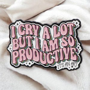 May include: A pink and white enamel pin with the text "I CRY A LOT BUT I AM SO PRODUCTIVE" in a retro font. The pin has a black border and the words "IT'S AN ART" at the bottom.