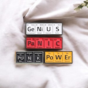 May include: Three rectangular magnets with the words "GENIUS", "PANIC", and "PUNK POWER" spelled out using periodic table elements. The magnets are black, red, and yellow with white lettering. The background is a white, textured fabric.