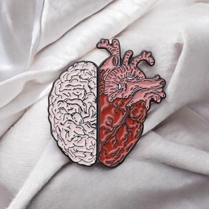 May include: A pin featuring a split design of a brain and a heart. The brain is light pink with detailed lines, while the heart is red and pink with intricate anatomical details. The pin is set against a white, textured background.