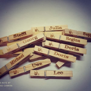 Könnte beinhalten: Holz-Wäscheklammern mit aufgedruckten Namen, darunter Steffi, Julia, Georg, Regina, Doris, Michael, Uwe, Leo und Sepp. Jede Wäscheklammer hat ein kleines, eingraviertes Bild von einem Eimer und einer Schaufel.