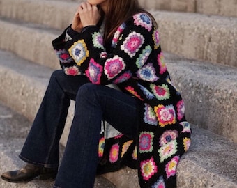 May include: A woman wearing a black crochet cardigan with colorful granny squares, blue jeans, and cowboy boots. She is sitting on a set of concrete steps.