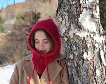 Può includere: Una donna indossa un cappello rosso lavorato a maglia con un collo lungo e lacci abbinati, in piedi davanti a un betulla. Indossa un cappotto color beige.