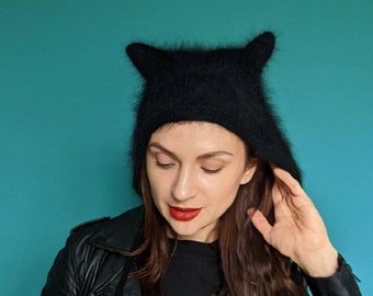 Può includere: Una donna indossa una giacca di pelle nera e un cappello nero peloso con orecchie da gatto. Sta guardando in basso e ha il rossetto rosso.