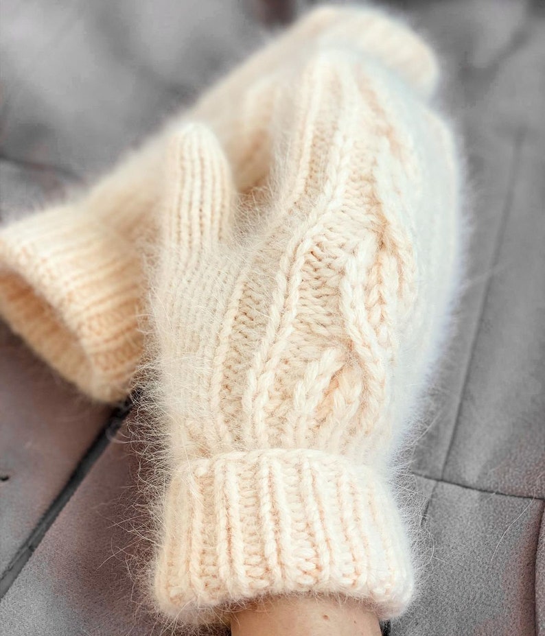 Pu&ograve; includere: Un paio di guanti morbidi di lana bianca con un motivo a treccia. I guanti sono realizzati con un filato soffice e hanno una texture pelosa.