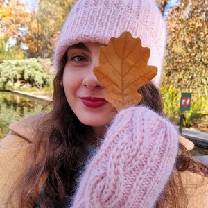 Pu&ograve; includere: Una donna indossa un cappello di lana rosa con un pon pon soffice, guanti di lana rosa e una giacca marrone. Sta tenendo una foglia marrone davanti al suo viso.