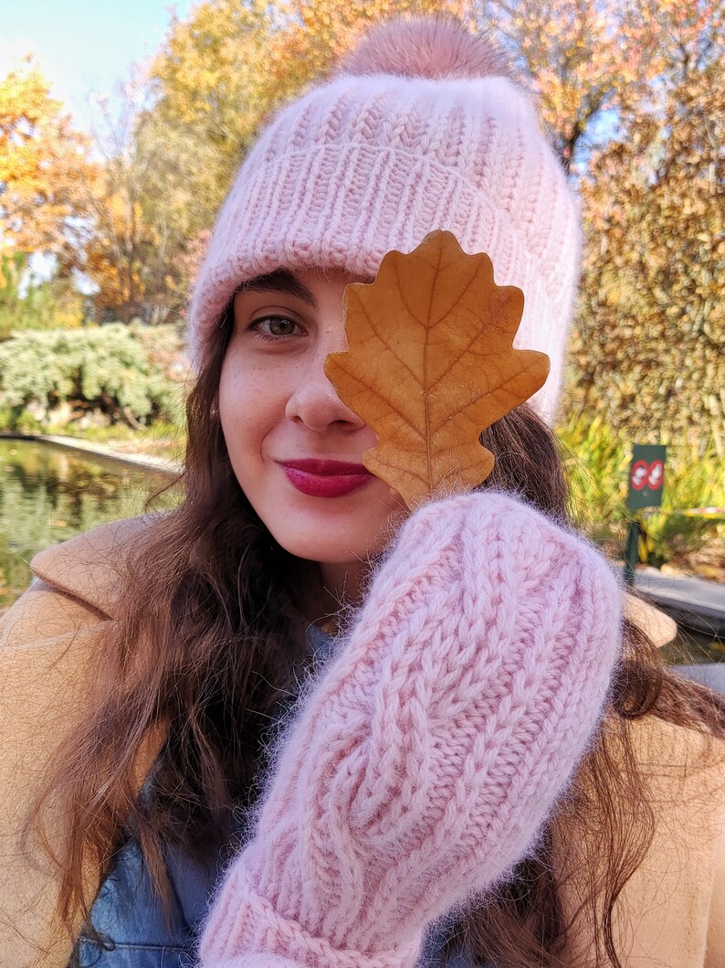 Pu&ograve; includere: Una donna indossa un cappello di lana rosa con un pon pon soffice, guanti di lana rosa e una giacca marrone. Sta tenendo una foglia marrone davanti al suo viso.